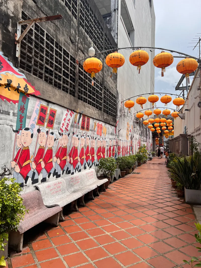 Narrow walkway with red-tiled flooring lined by potted plants and stone benches, bordered by a long mural depicting rows of cartoon-style figures in red clothing holding staffs. Overhead, multiple bright orange lanterns hang in a row from metal arches. The alley is framed by tall, weathered buildings on both sides, with a few people visible in the distance.