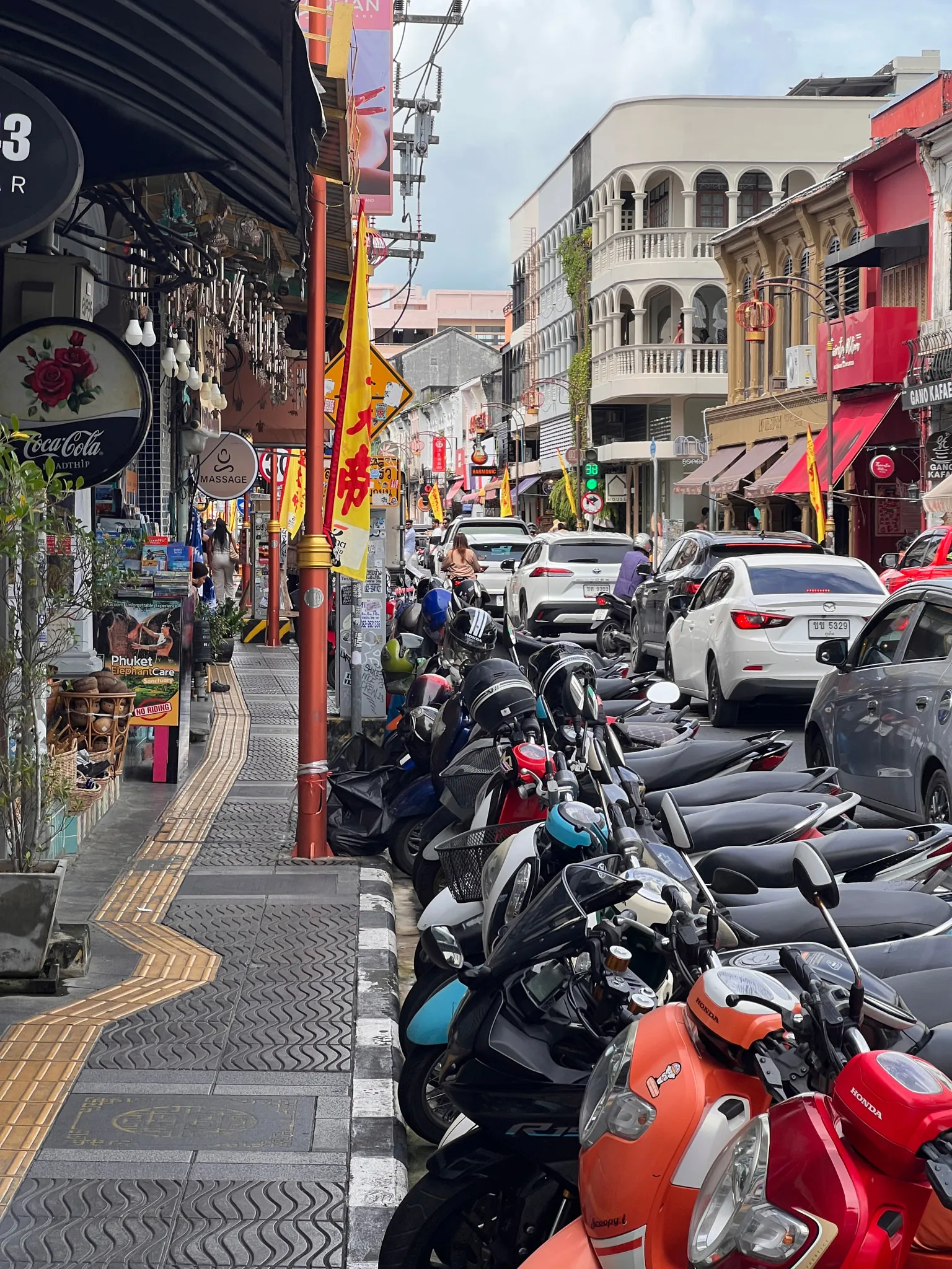 A lively urban street filled with parked motorbikes along the curb and slow‑moving traffic of cars and scooters. Storefronts line both sides of the street, featuring colorful signs, awnings, and decorative elements. Yellow festival or event flags hang from poles, adding bright accents to the scene. Pedestrians walk along a narrow patterned sidewalk with tactile paving, while overhead wires and shop lights create a dense, busy atmosphere. The architecture includes a mix of older and modern buildings with balconies and detailed facades.