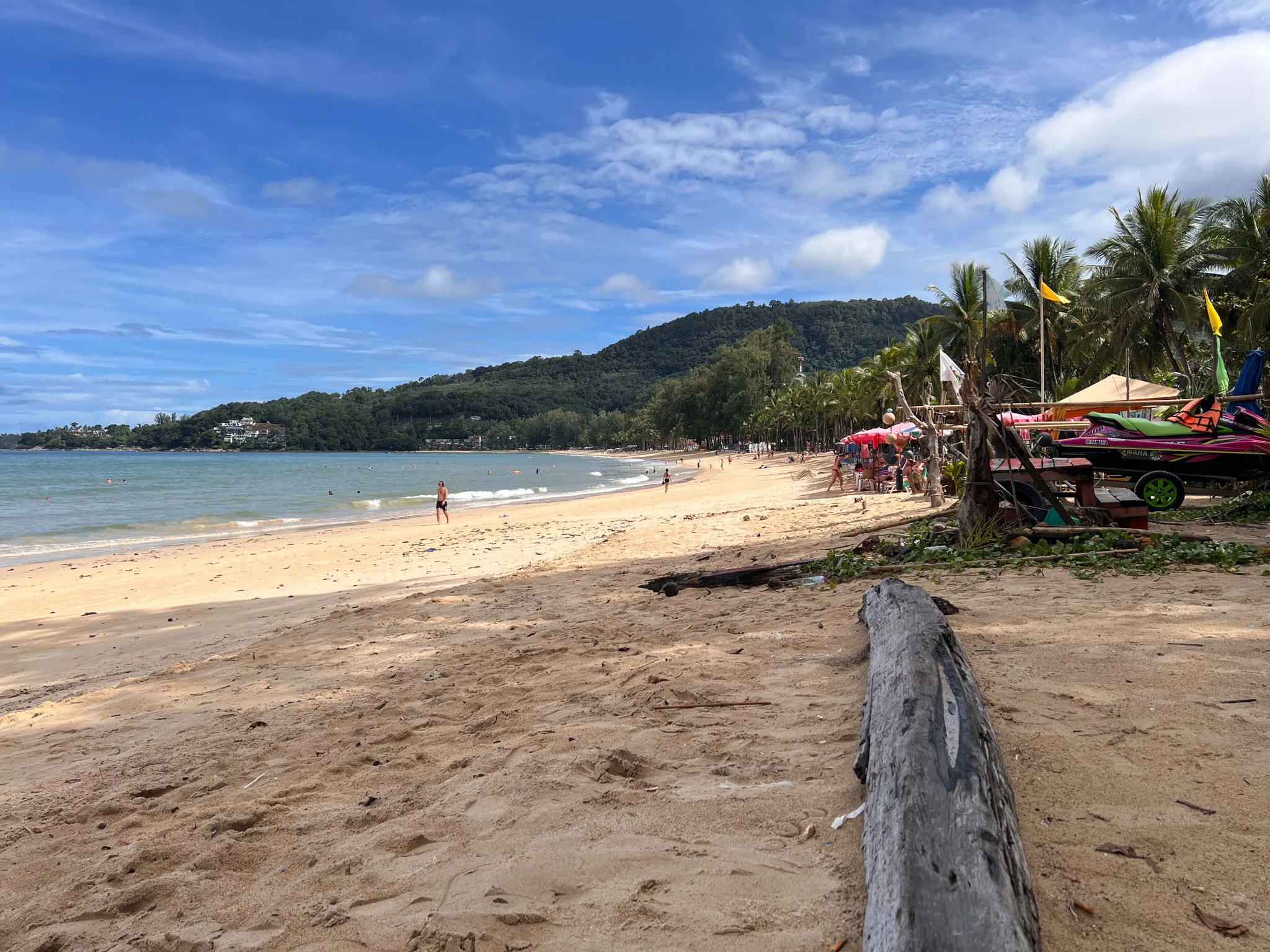 Sandy beach stretching along calm blue-green water with small waves approaching the shore. A few people walk near the water while others swim farther out. A long piece of driftwood lies in the foreground on the sand. To the right, colorful umbrellas, palm trees, and watercraft create a lively area near the edge of the beach. Green hills rise in the background beneath a bright sky filled with scattered clouds.