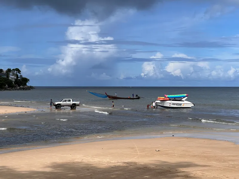 A sandy beach stretches toward shallow coastal water where several people stand near two boats and a white pickup truck. One long wooden boat with a blue tarp is anchored further out, while a white boat carrying colorful inflatable tubes is positioned closer to shore. Gentle waves roll in under a partly cloudy sky, and a small tree-covered rocky area is visible on the left in the distance.