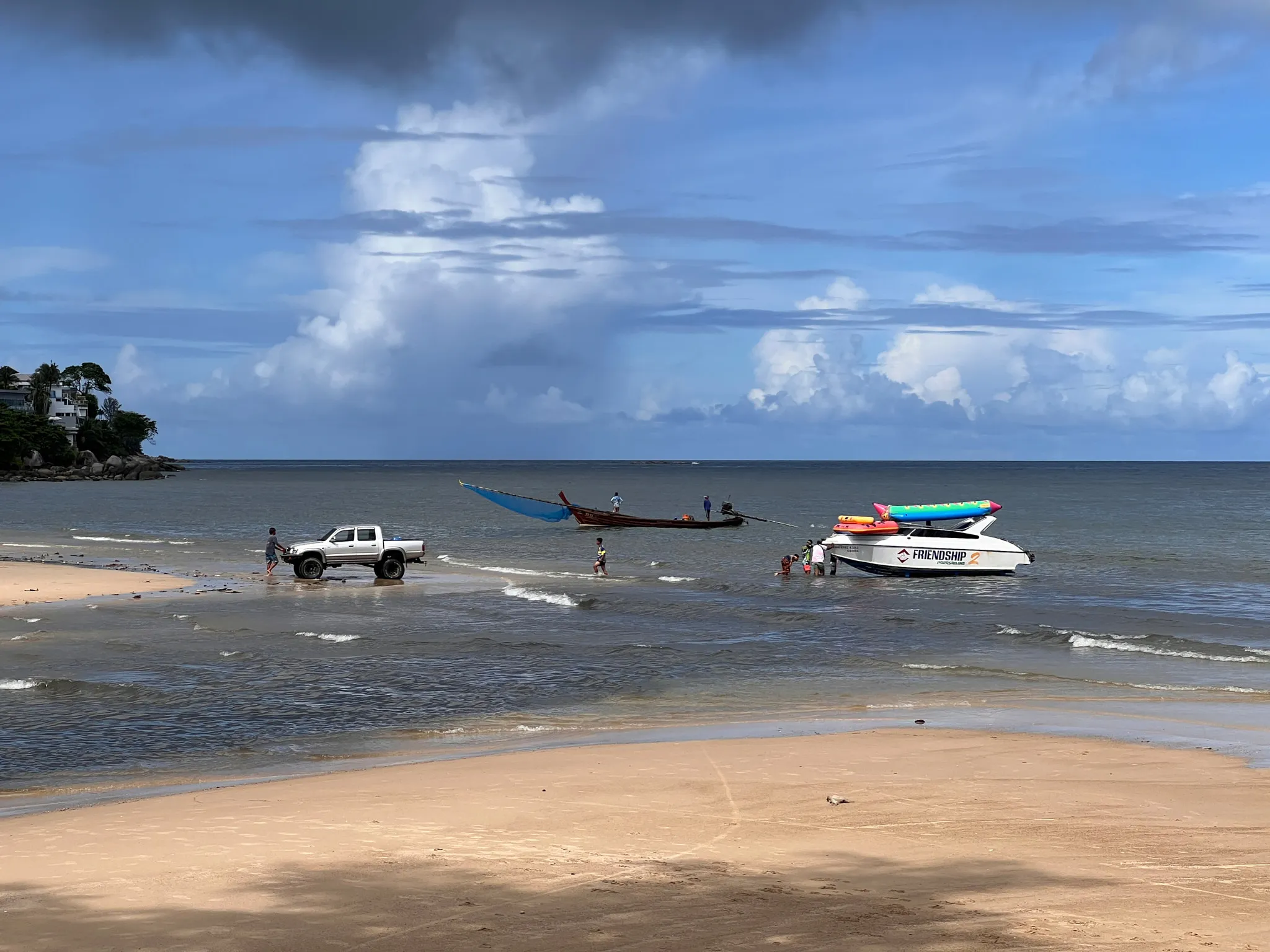 A sandy beach stretches toward shallow coastal water where several people stand near two boats and a white pickup truck. One long wooden boat with a blue tarp is anchored further out, while a white boat carrying colorful inflatable tubes is positioned closer to shore. Gentle waves roll in under a partly cloudy sky, and a small tree-covered rocky area is visible on the left in the distance.