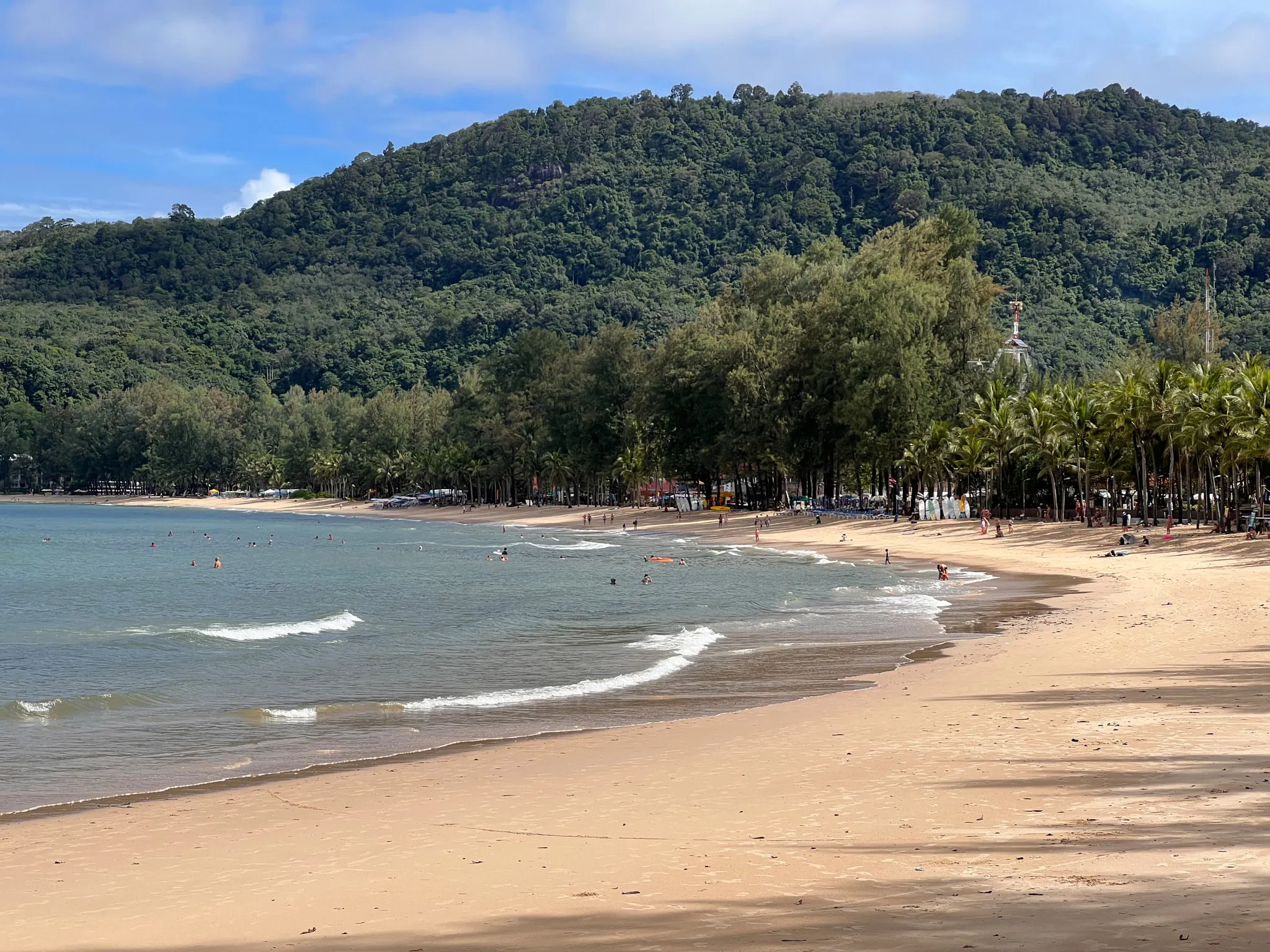 A wide sandy beach curves gently along a calm bay with small rolling waves approaching the shore. Several people swim and relax in the shallow water, while others stroll or sit along the beach. Dense green hills rise in the background, covered in lush forest. A line of tall trees and palms borders the back of the beach, providing shade and separating the sand from nearby structures. The sky is lightly clouded with patches of blue, creating a peaceful tropical atmosphere.