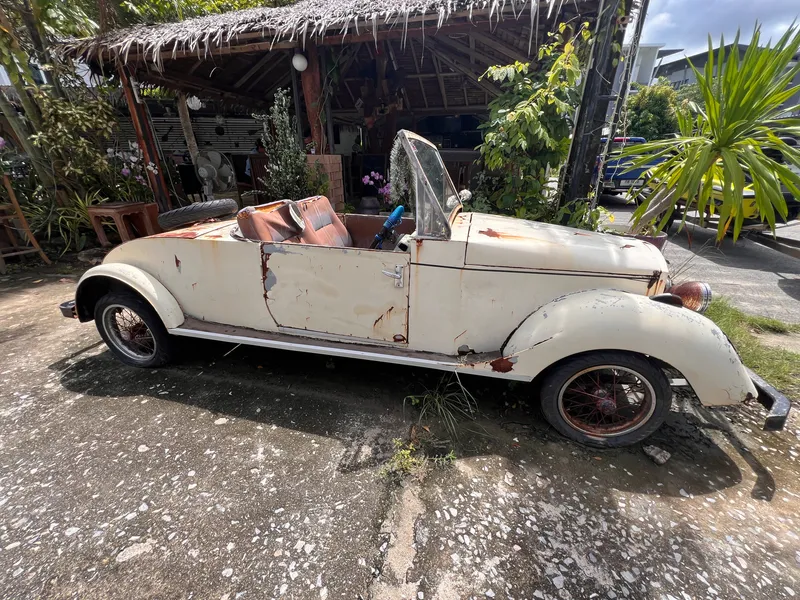 A weathered cream-colored convertible with significant rust and peeling paint sits parked on a rough concrete surface beside a rustic, thatched-roof structure. The car’s interior is worn, with faded brown seats and a missing or damaged roof. Overgrown plants and tropical greenery surround the area, while scattered tools and objects rest under the open wooden shelter. Sunlight filters through the environment, highlighting the aged condition of the vehicle and the relaxed, outdoor setting.