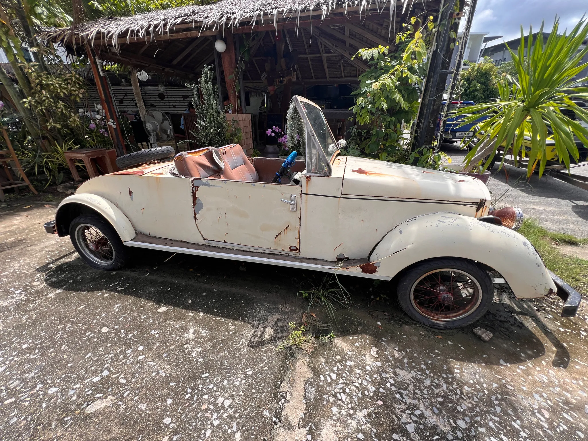 A weathered cream-colored convertible with significant rust and peeling paint sits parked on a rough concrete surface beside a rustic, thatched-roof structure. The car’s interior is worn, with faded brown seats and a missing or damaged roof. Overgrown plants and tropical greenery surround the area, while scattered tools and objects rest under the open wooden shelter. Sunlight filters through the environment, highlighting the aged condition of the vehicle and the relaxed, outdoor setting.