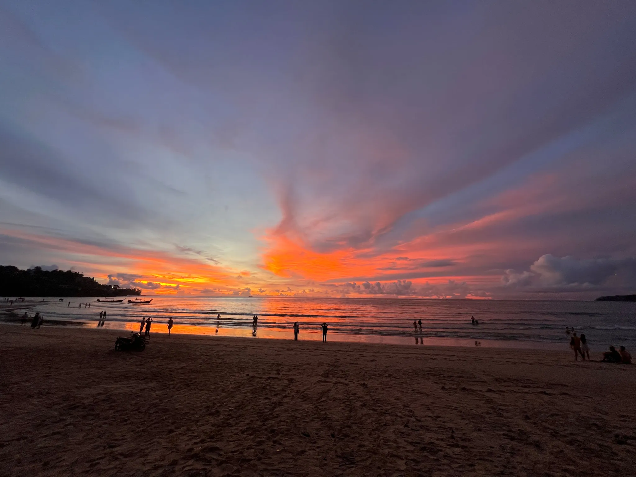 A vivid sunset spreads across the sky with deep oranges, pinks, and purples blending into the darker blue tones above. Gentle waves roll onto the shore, reflecting the warm colors of the sky on the wet sand. Silhouetted figures stand, walk, and play along the water’s edge, while small boats rest near the horizon. Wisps of clouds stretch outward from the glowing center of the sunset, adding texture to the expansive sky. The beach appears calm and spacious, with people scattered peacefully across the scene.