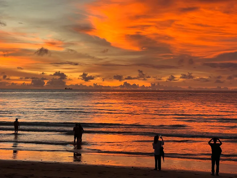 A vivid sunset fills the sky with deep orange and red tones above a calm ocean. Silhouetted figures stand along the shoreline, some wading in the shallow water and others watching or photographing the dramatic sky. Gentle waves roll toward the beach, reflecting the intense colors of the sunset, while dark clouds scatter across the horizon.