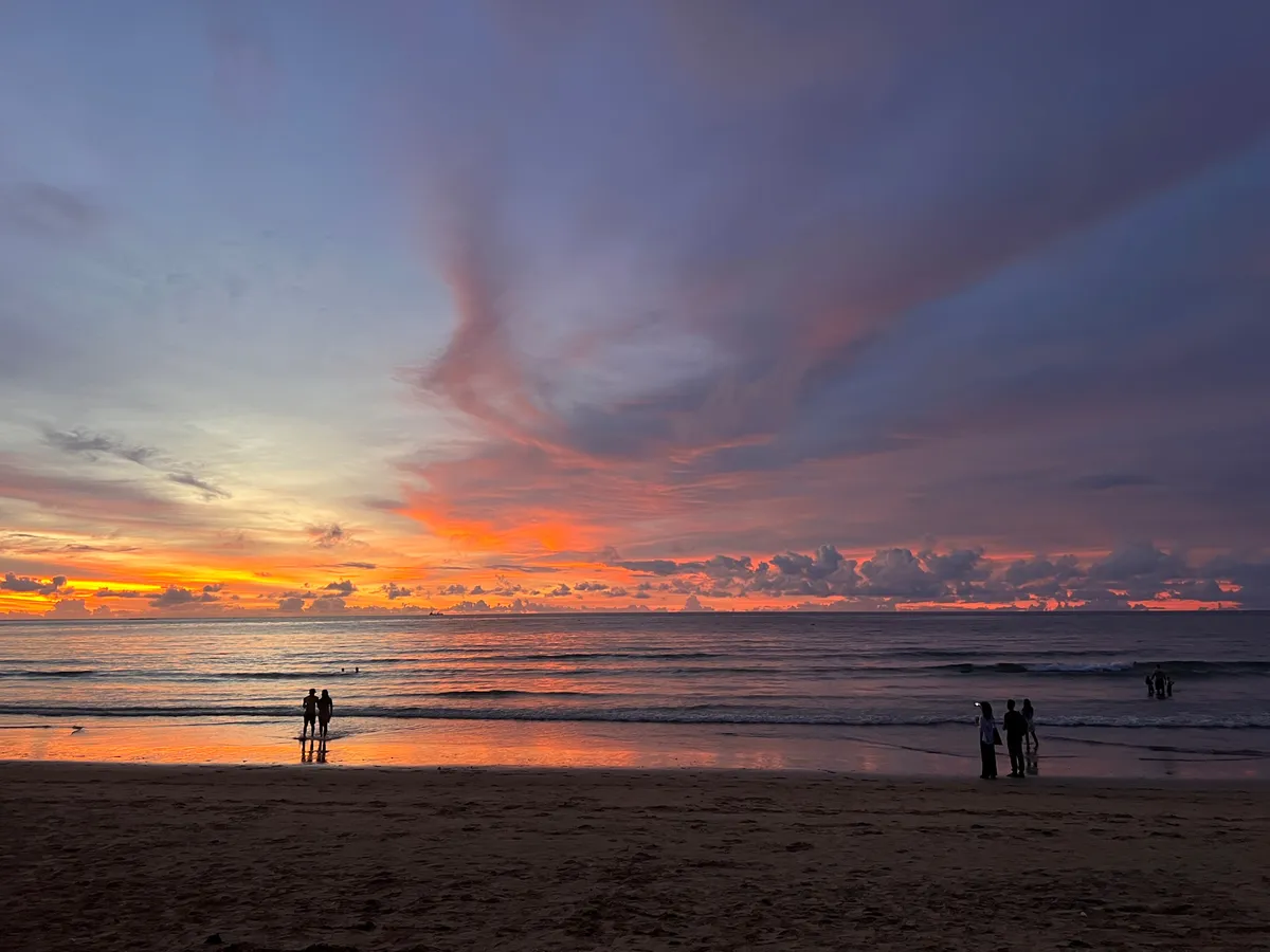 Spectacular sunset at Kamala Beach in Phuket Province, Thailand. The sky glows in a dramatic play of colors — luminous orange, soft pink, deep violet, and gentle blue — with the colors spreading in broad cloud formations across the entire sky. On the horizon, the sun disappears behind cloud banks, bathing the sky in intense orange tones that reflect on the wet sand surface and the calm water of the Andaman Sea. In the foreground extends the broad sandy beach of Kamala, on which several silhouettes of visitors can be seen — a couple stands at the waterline on the left, while a small group on the right takes photos with a smartphone. Gentle waves roll onto the beach, and the reflections of the colorful sky give the wet sand a warm, golden shimmer.