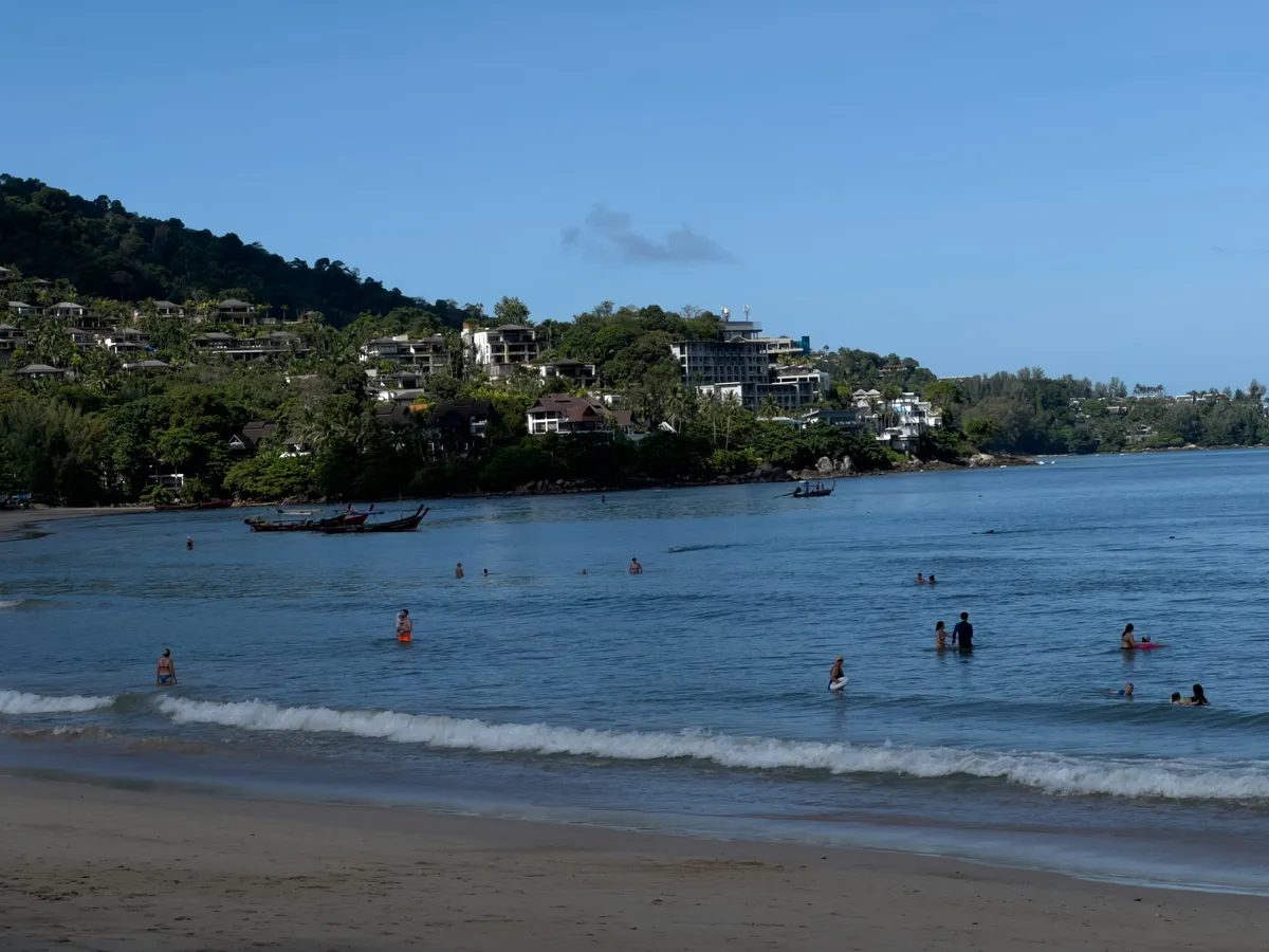 View across the beach of Kamala in Phuket Province, Thailand. In the foreground extends a sandy beach with gently rolling waves forming white foam. In the calm, deep-blue sea, numerous bathers wade and swim in the shallow water. On the left side, traditional Thai longtail boats are moored near the shore. In the background rises a lushly green hill, on whose slopes several resorts, hotels, and villas are nestled into the tropical vegetation. The sky is largely clear with occasional small clouds, indicating a sunny day. The bay of Kamala offers a picturesque backdrop with the combination of turquoise-blue sea, tropical vegetation, and the buildings on the hillside.